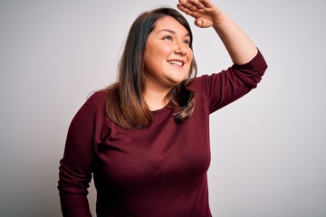 Beautiful brunette plus size woman wearing casual sweater over isolated white background very happy and smiling looking far away with hand over head. Searching concept.