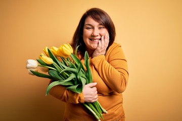 Beautiful plus size woman holding romantic bouquet of natural tulips flowers over yellow background laughing and embarrassed giggle covering mouth with hands, gossip and scandal concept