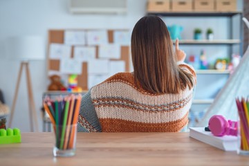 Young beautiful teacher woman wearing sweater and glasses sitting on desk at kindergarten Posing backwards pointing ahead with finger hand