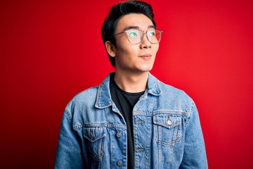 Young handsome chinese man wearing denim jacket and glasses over red background smiling looking to the side and staring away thinking.