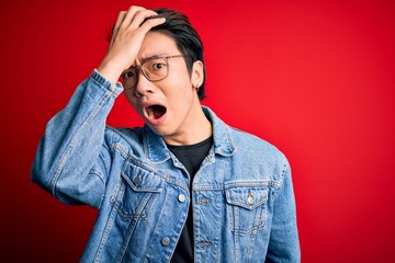 Young handsome chinese man wearing denim jacket and glasses over red background surprised with hand...