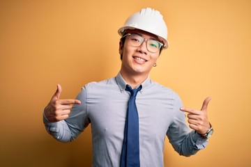 Young handsome chinese architect man wearing safety helmet and tie over yellow background looking confident with smile on face, pointing oneself with fingers proud and happy.