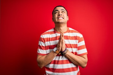 Young brazilian man wearing casual striped t-shirt standing over isolated red background begging and praying with hands together with hope expression on face very emotional and worried. Begging.