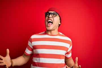 Young brazilian man wearing casual striped t-shirt and glasses over isolated red background crazy and mad shouting and yelling with aggressive expression and arms raised. Frustration concept.