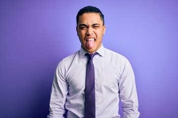 Young brazilian businessman wearing elegant tie standing over isolated purple background sticking tongue out happy with funny expression. Emotion concept.