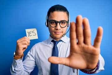 Young brazilian call center agent man holding reminder paper with yes message with open hand doing...