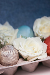 Fresh white hybrid tea rose flowers and baby's breath (Gypsophila) with natural-dyed eggs with botanical leaf prints displayed in an egg carton against a denim blue background, with copy space