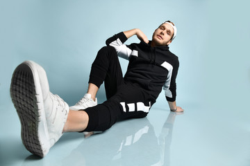 Hipster person in cap, black tracksuit, bracelet and sneakers. He is sitting on floor, posing against blue background. Close up