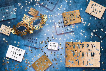 Happy Purim written in light box, carnival mask and golden confetti on blue background. Flat lay of Purim Carnival celebration concept.