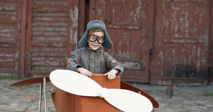 Caucasian cute funny little boy with red hair in hat and glasses sitting outdoor in wooden toy of plane and walking the yard. Happy kid dreaming to be pilot when grow up.