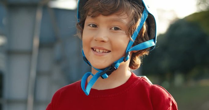 Close up of cute Caucasian small boy in helmet and red sweater standing outdoor on sunny day at big bridge. Portrait of little kid with smile at bike riding. Childhood activity concept.