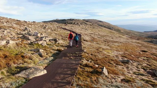 Evening scenic aerial drone view of a young, sporty hiker couple walking over the rusty steel boardwalk near the summit of Mount Kosciuszko in Kosciuszko National Park, New South Wales, Australia.