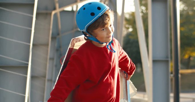 Cute Caucasian little boy in helmet and red sweater with carton wings of plane standing and dancing outdoors. Little kid with smile playing in airplane game. Childhood olay activity concept.