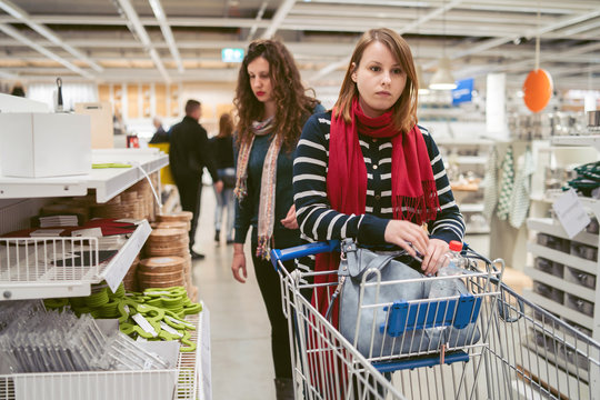 Two Beautiful Caucasian Women Girls Friends Walking By The Shelf At The Shopping Mall Pushing The Wire Metal Shopping Cart Checking The Products And Price Looking Confused Or Scared Mad Disappointed