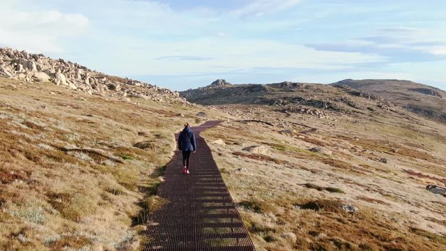 Evening scenic aerial drone view of a young, sporty female hiker walking over the rusty steel boardwalk near the summit of Mount Kosciuszko in Kosciuszko National Park, New South Wales, Australia.