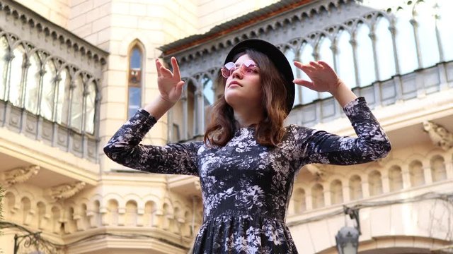 4K video cute stylish young girl in pink glasses with a light dress and a black hat on the background of an old courtyard hugs an evergreen plant with one hand