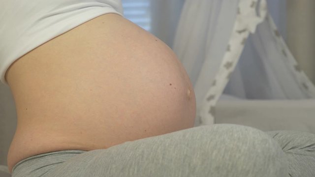 Baby moves in the mother's stomach in close-up. Pregnant woman sits on the bed and listens to unborn baby kicking in her belly. Young woman in last month of pregnancy.