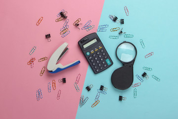 Office tools, stationery. Stapler, calculator, magnifier, paper clips on a blue-pink pastel...