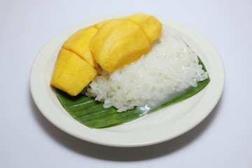 Most popular Thai  dessert, ripe yellow mango with sticky rice served with coconut milk  on banana leaf on white plate