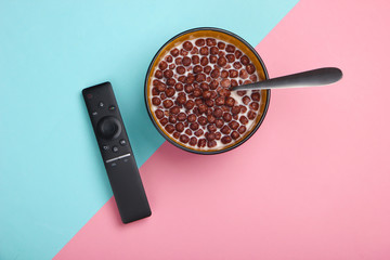 TV remote control and chocolate cereal balls with milk in bowl on pink blue background. Watching the TV. The concept of home rest after work. Top view.