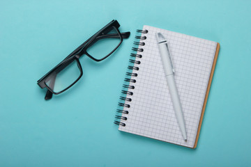 Classic eyeglasses and notebook with pen on a blue pastel background. Top view