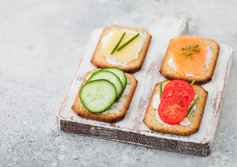 Various healthy crackers with salmon and cheese, tomato and cucumber on wooden chopping board on light kitchen table background. Space for text