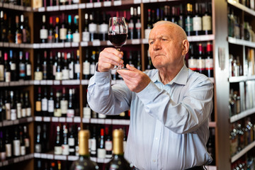 Older man inspecting quality of red wine in wine store in search of perfect wine for solemn occasion