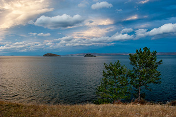 View of the lake with two trees in the foreground. Water with ripples. Two islands on the lake. Clouds in the sky. Sunset glare.