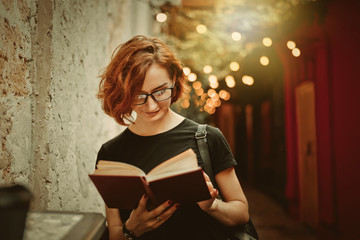 Young hipster woman in glasses with short haircuts reading book in outdoor street cafe