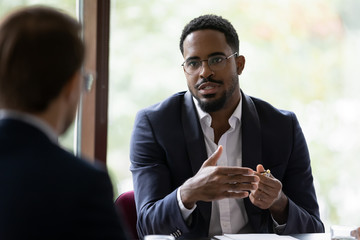 Confident concentrated African American male employee talk with colleague explain thought or idea, focused biracial businessman speak with coworker or partner, brainstorm at office boardroom meeting - obrazy, fototapety, plakaty