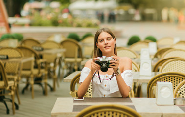 Young keen woman holds old retro camera in her hands while sitting at the table in outdoor cafe