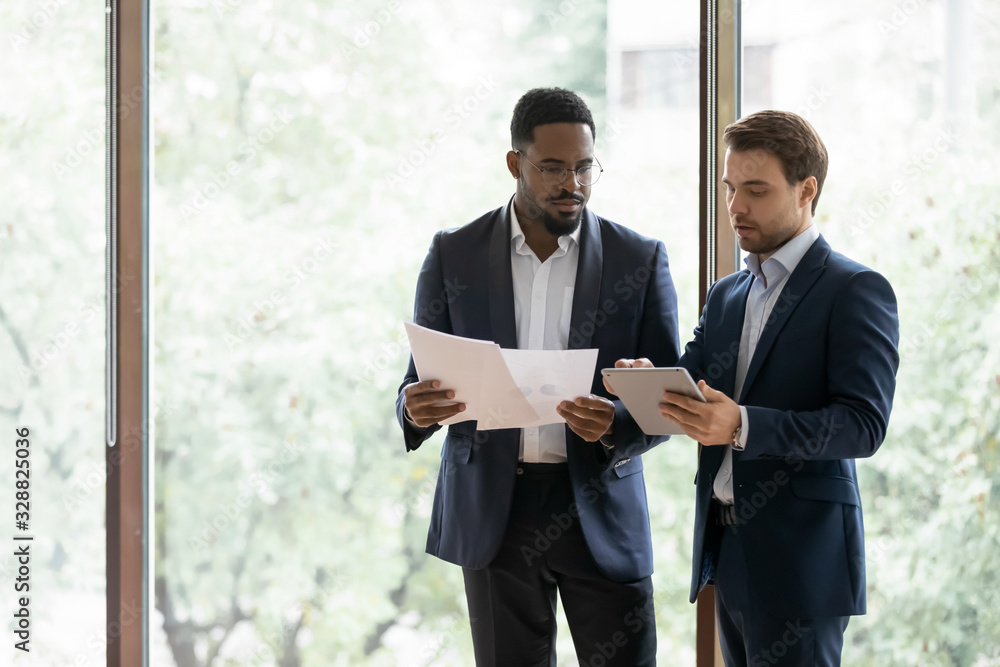 Sticker concentrated diverse businessmen stand near window brainstorm discuss company paperwork together, se