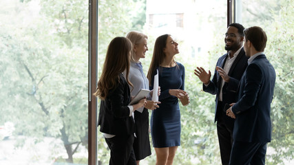 Smiling multiracial colleagues stand talking having fun during company work break in office, overjoyed diverse multiethnic employees speak joke discuss ideas or project near big window in boardroom