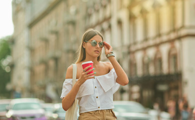 Fototapeta premium Coffee on the go. Young fashion woman in trendy summer clothes holding coffee cup while walking around the city and posing for the camera