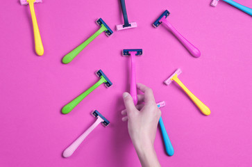 Female hand choosing green razor among a variety of colored epilator razors on pink background. Top view. Minimalism beauty concept. Top view