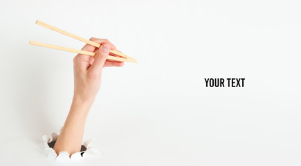 Female hand hold chopsticks through torn hole on white background. Minimalistic food concept. Copy space. Top view