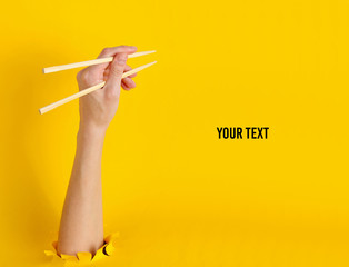 Female hand hold chopsticks through torn hole on yellow background. Minimalistic food concept. Copy space. Top view