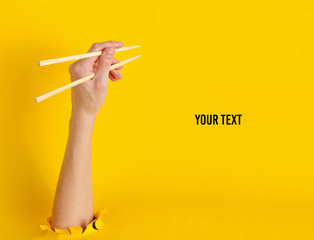 Female hand hold chopsticks through torn hole on yellow background. Minimalistic food concept. Copy...