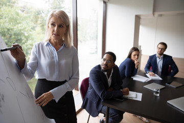 Focused middle-aged confident businesswoman make flip chart presentation for diverse coworkers,...