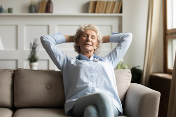 Calm senior 60s woman sit relax on comfortable sofa in living room taking nap or daydreaming at...