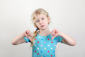 Unhappy preschool child kid showing dislike sign thumb fingers down. Portrait of little sad upset blonde Caucasian girl on light background in a studio. Negative emotion face expression.