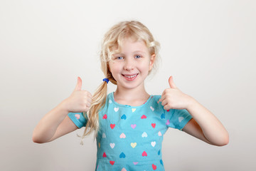 Portrait of beautiful little funny joyful blonde Caucasian girl on a light background. Happy smiling preschool child kid showing like thumb fingers. Positive emotion face expression.