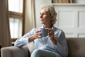 Pensive senior 60s woman sit rest on couch in living room drink tea or coffee look in distance...