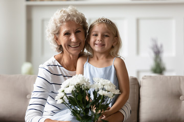 Family portrait of cute little granddaughter congratulating smiling senior grandmother with birthday or anniversary, loving small grandchild make surprise greeting excited granny presenting flowers