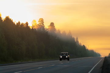 Passenger car drives on foggy highway in morning forest, sunrise orange sky with empty road