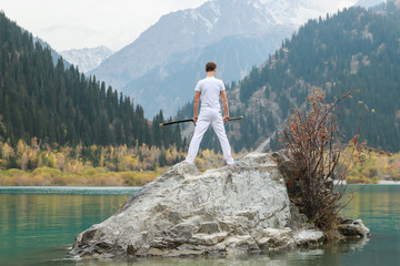 A man in white clothes with a sword stands on a large stone among the highlands.