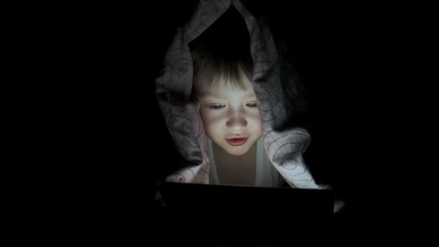 little boy plays on tablet while lying under blanket. watching cartoons hiding