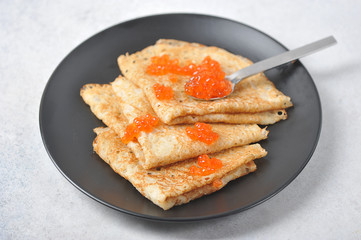 Pancakes on a black plate. On pancakes, red caviar and a spoon with red caviar. Close-up. View from above.