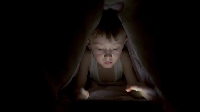 little boy plays on tablet while lying under blanket. watching cartoons hiding
