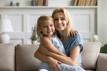 Portrait of smiling young mother and cute little preschooler daughter sit on couch in living room look at camera, happy mom and small girl child hug embrace relax on sofa at home, bonding concept